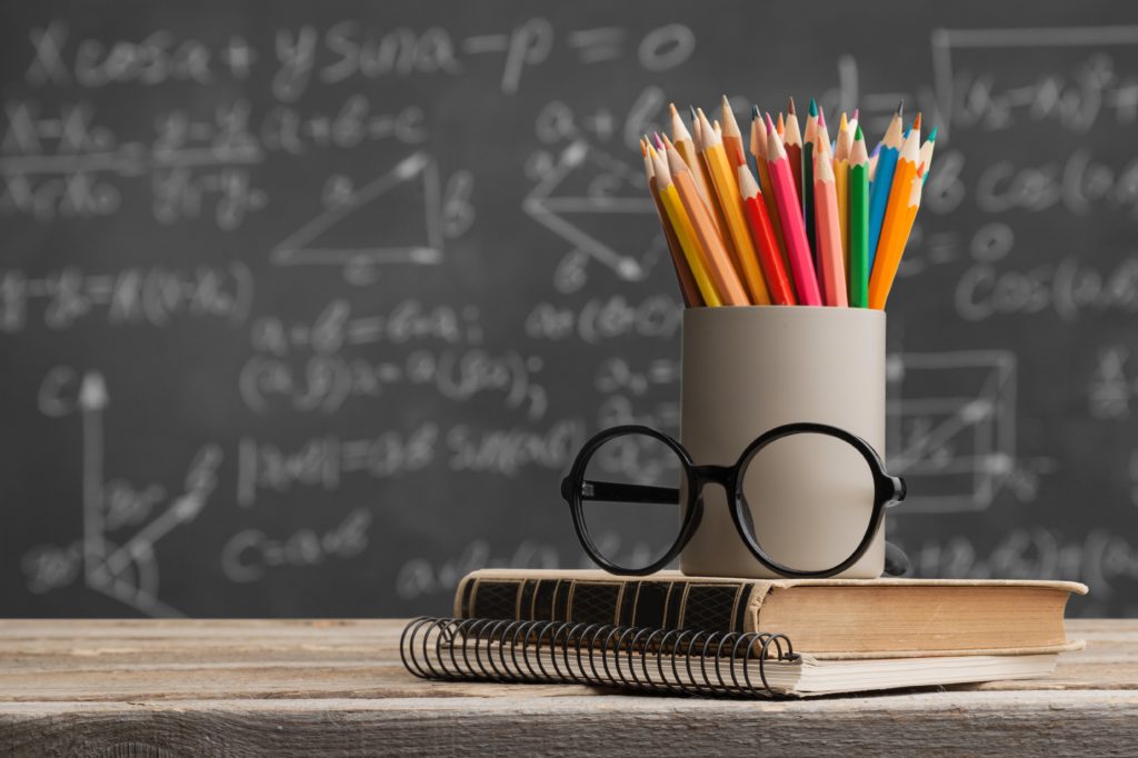 Colored pencils and glasses sit on a book and notebook in front of a blackboard.