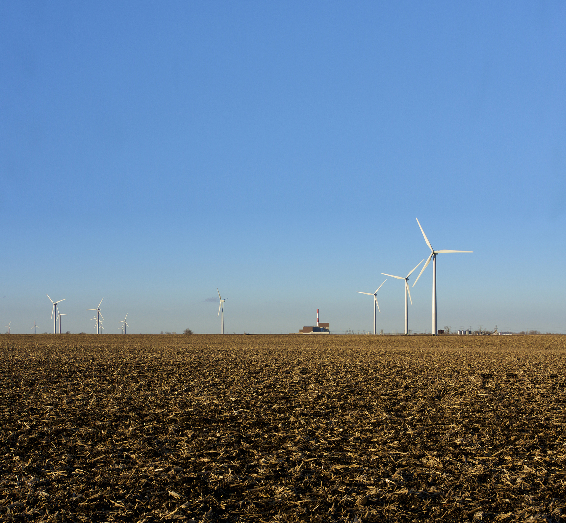 Photo of windmill towers in a farm field with a power plant in the background